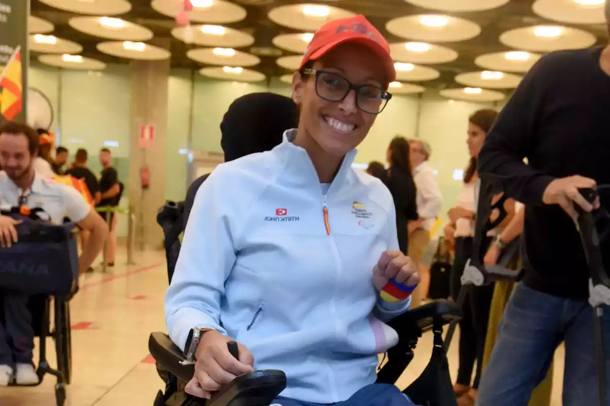 Una mujer en silla de ruedas, vestida con una chaqueta deportiva blanca y una gorra roja, sonríe mientras está en un lugar público con varias personas alrededor.