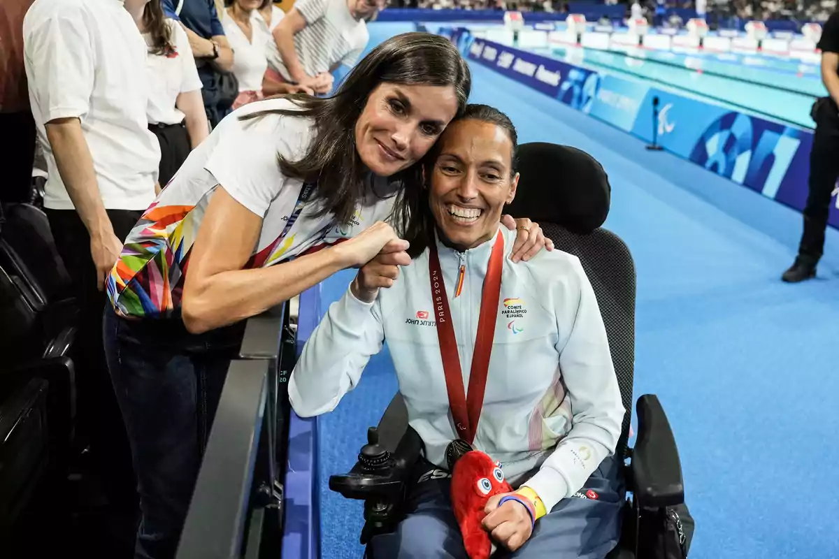 Una mujer se inclina para abrazar y felicitar a una atleta en silla de ruedas que lleva una medalla y un peluche, mientras están en un evento deportivo con una piscina de fondo.