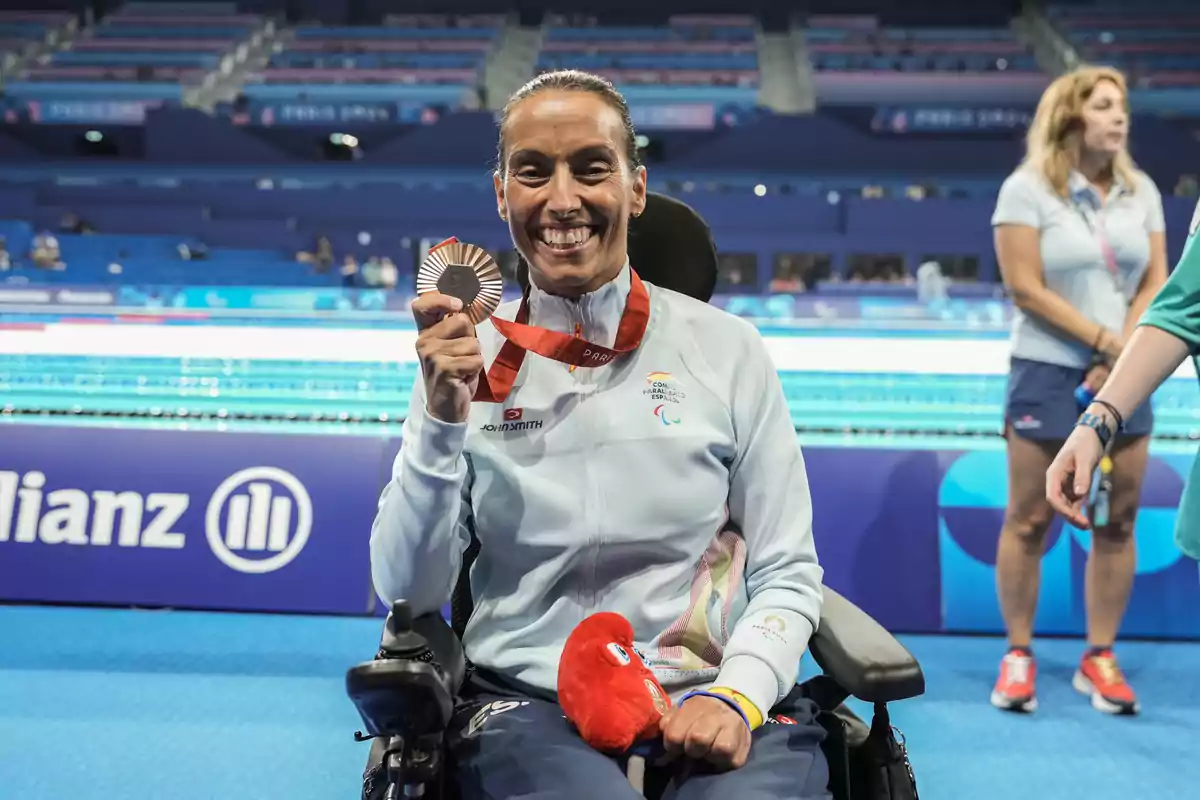 Una atleta en silla de ruedas sonríe mientras sostiene una medalla frente a una piscina, con personas y gradas en el fondo.