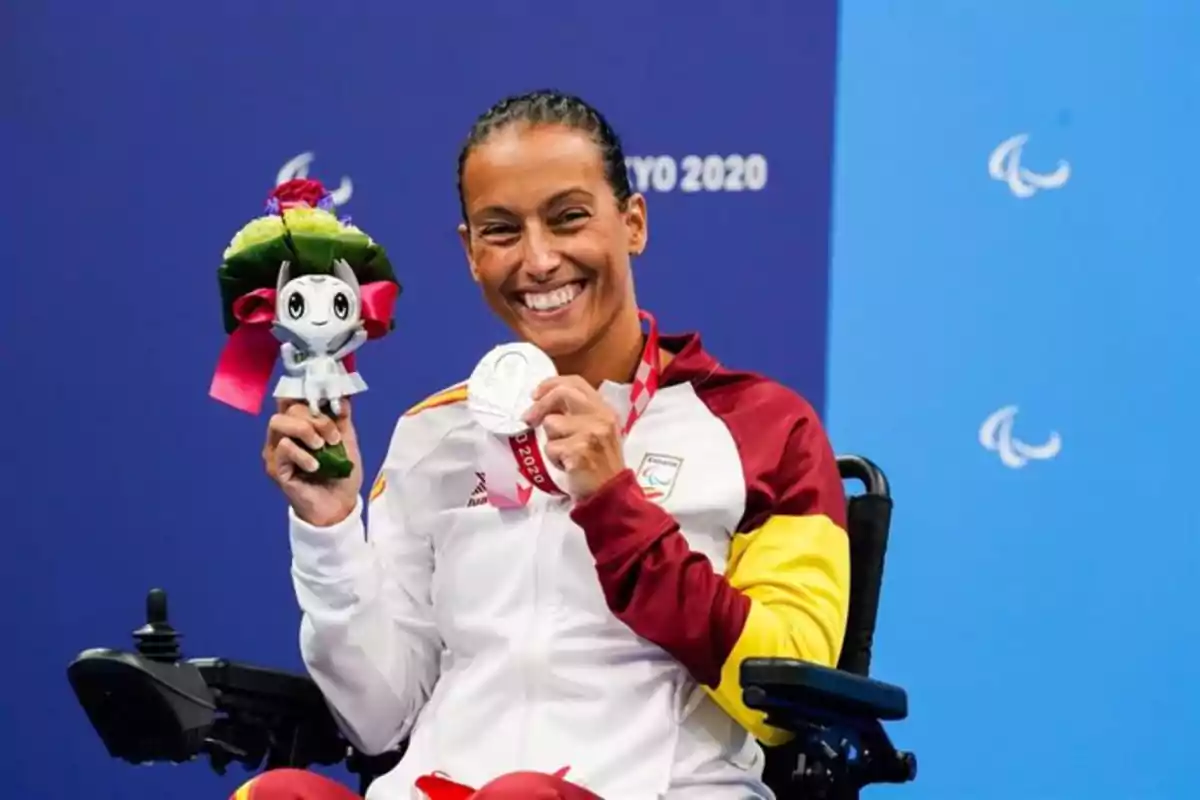 Persona en silla de ruedas sonriendo y mostrando una medalla y una mascota de los Juegos Paralímpicos de Tokio 2020.