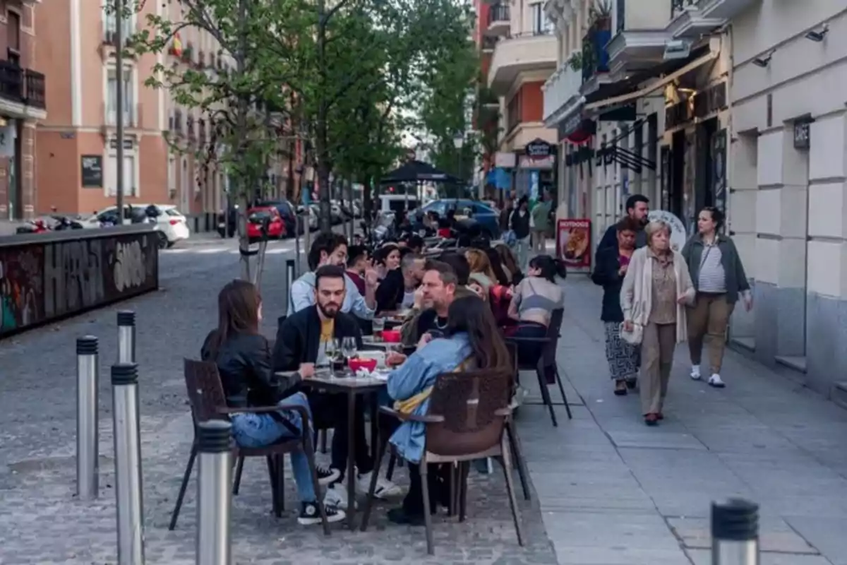 Personas sentadas en mesas al aire libre en una terraza de una calle urbana mientras otras personas caminan por la acera Personas sentadas en mesas al aire libre en una terraza de una calle urbana mientras otras personas caminan por la acera