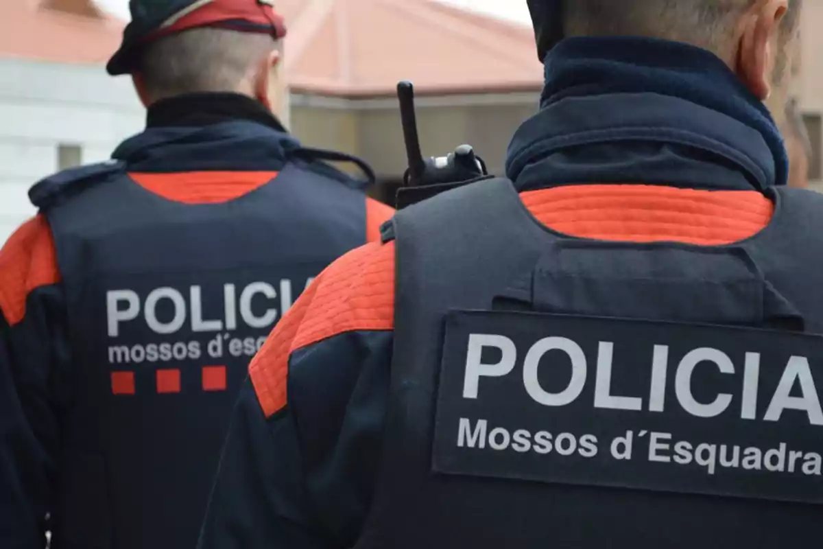 Dos agentes de los Mossos d'Esquadra de espaldas con uniformes oscuros y detalles rojos.