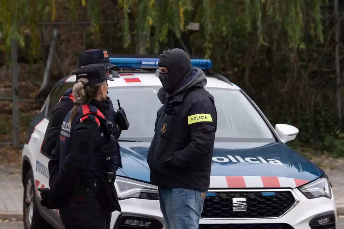 Dos agentes de policía conversan con un hombre enmascarado frente a un coche patrulla.