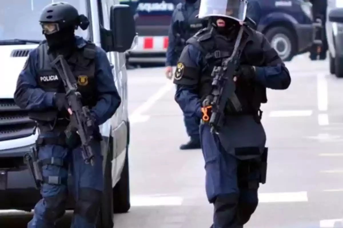 Dos policías con equipo táctico y armas caminan junto a una furgoneta en una calle.