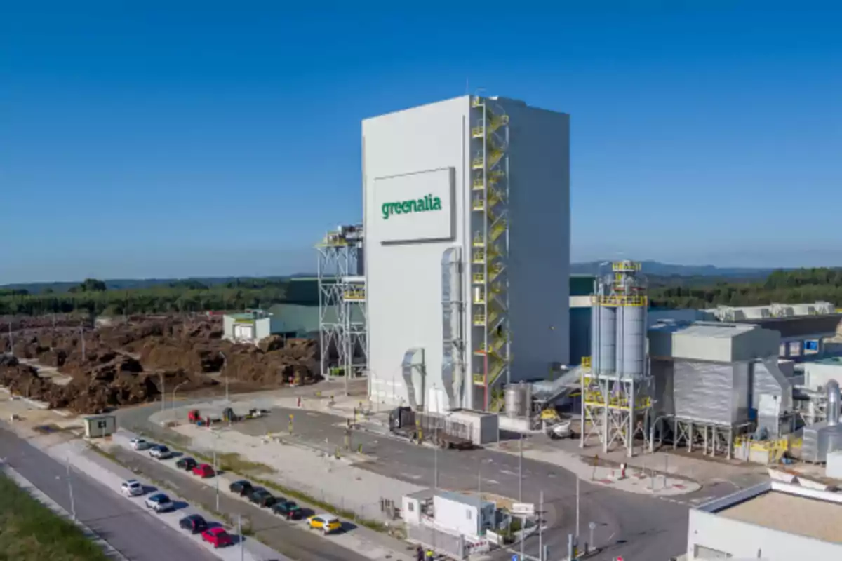 Vista aérea de una planta industrial de biomasa con el logo de Greenalia en el edificio principal rodeada de pilas de material orgánico y varios automóviles estacionados Vista aérea de una planta industrial de biomasa con el logo de Greenalia en el edificio principal rodeada de pilas de material orgánico y varios automóviles estacionados