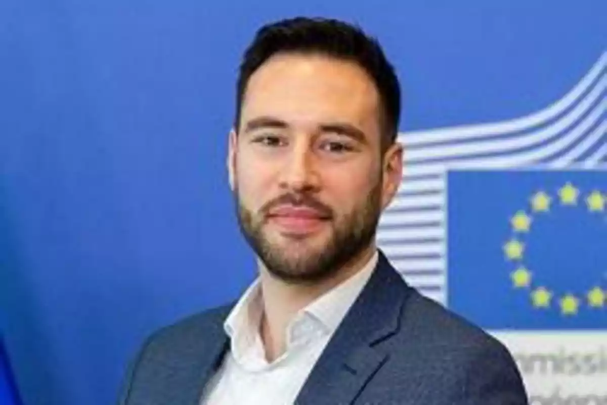 Hombre con barba y traje frente a un fondo azul con el logo de la Unión Europea.