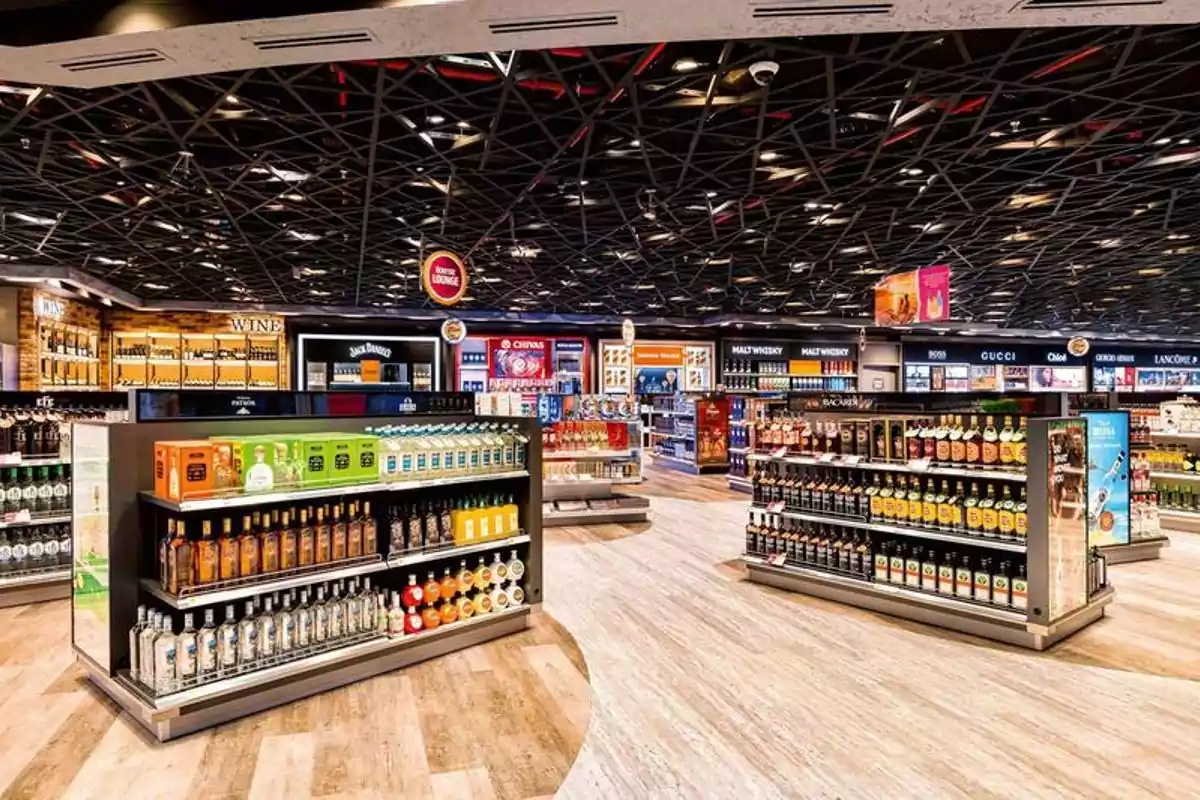 Una tienda de licores bien iluminada con estantes llenos de botellas de diferentes marcas y tipos de bebidas alcohólicas, con un techo de diseño moderno y un suelo de madera clara.