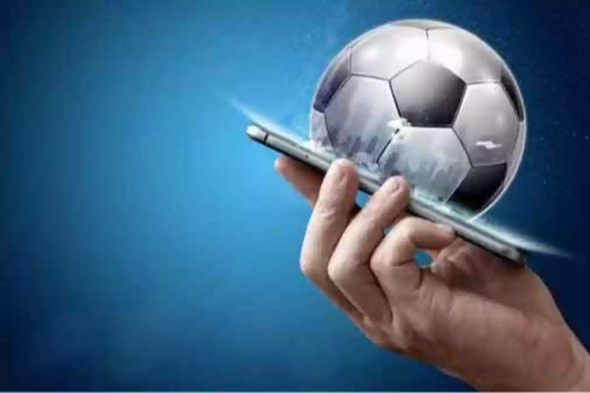 Mano sosteniendo un teléfono móvil con un balón de fútbol saliendo de la pantalla.