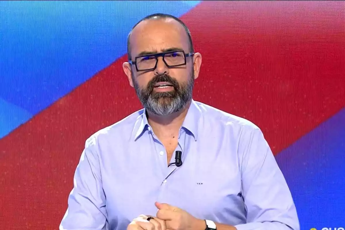 Un hombre con barba y gafas está hablando en un programa de televisión con un fondo colorido.