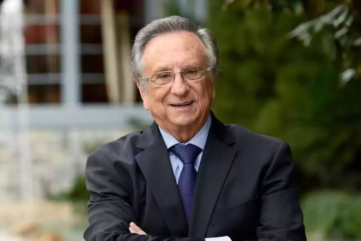 Un hombre mayor con gafas y traje sonríe al aire libre.