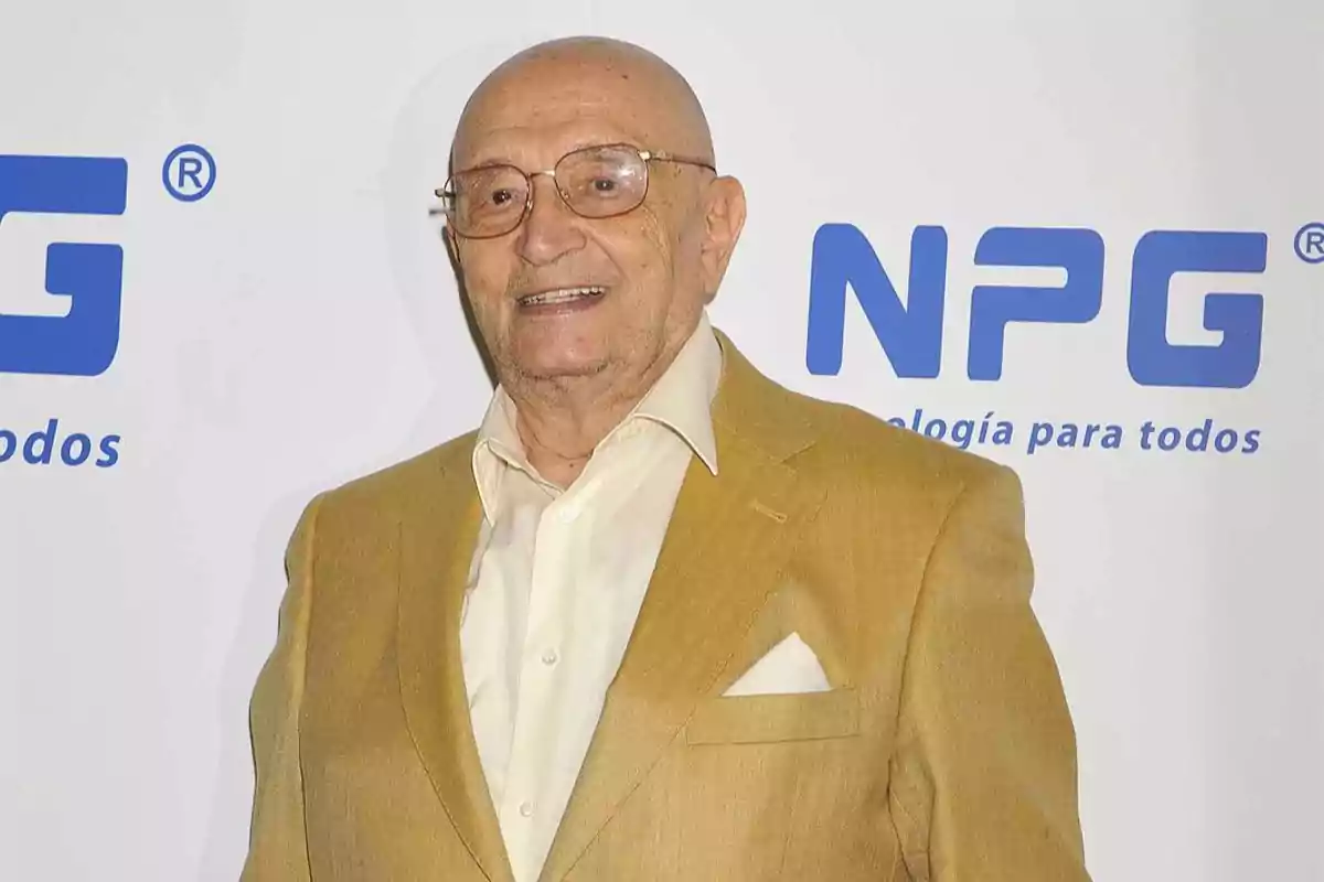 Un hombre mayor con gafas y traje beige sonríe frente a un fondo con el logo de NPG.