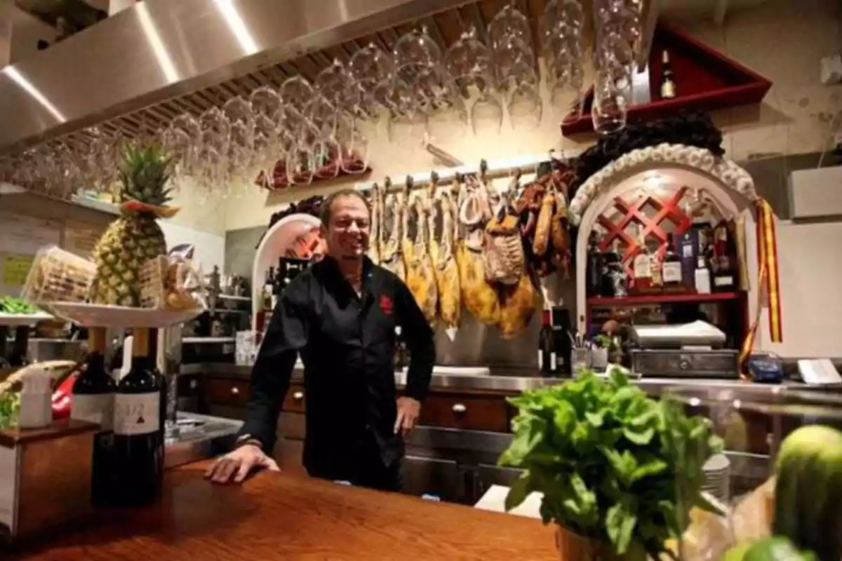 Un hombre sonriente detrás de la barra de un restaurante con jamones colgados y copas de vino invertidas sobre él.