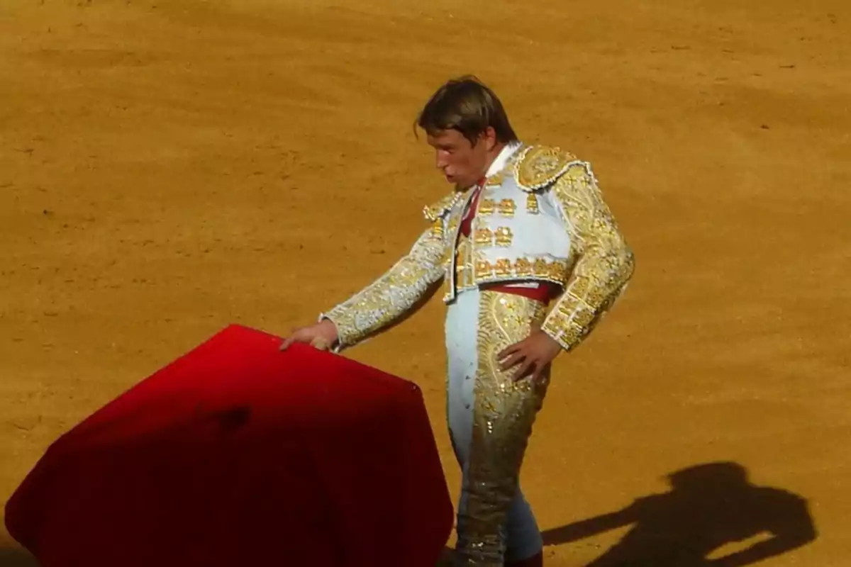 Un torero vestido con traje de luces dorado y blanco sostiene una muleta roja en una plaza de toros.