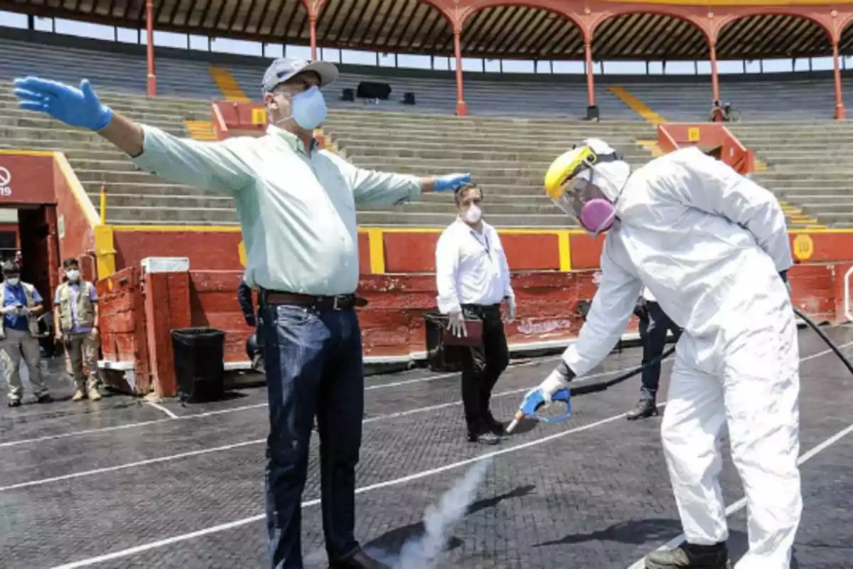 Un hombre con mascarilla y guantes es desinfectado por una persona con traje protector en un lugar público con gradas. Un hombre con mascarilla y guantes es desinfectado por una persona con traje protector en un lugar público con gradas.