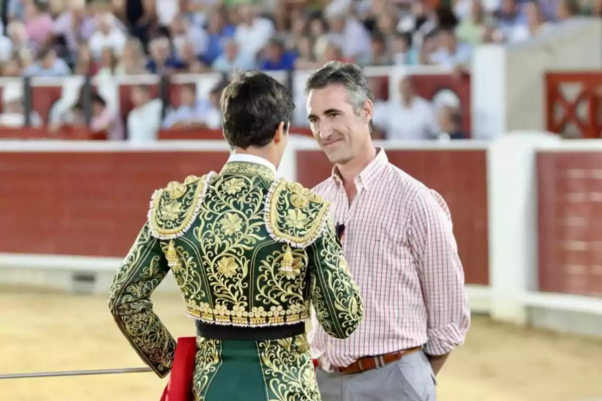 Un torero con traje verde y dorado conversa con un hombre de camisa a cuadros en una plaza de toros llena de espectadores.