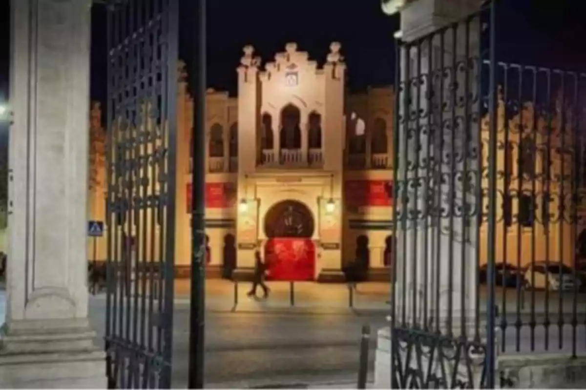 Entrada principal de un edificio histórico iluminado por la noche, vista a través de una reja de hierro forjado. Entrada principal de un edificio histórico iluminado por la noche, vista a través de una reja de hierro forjado.