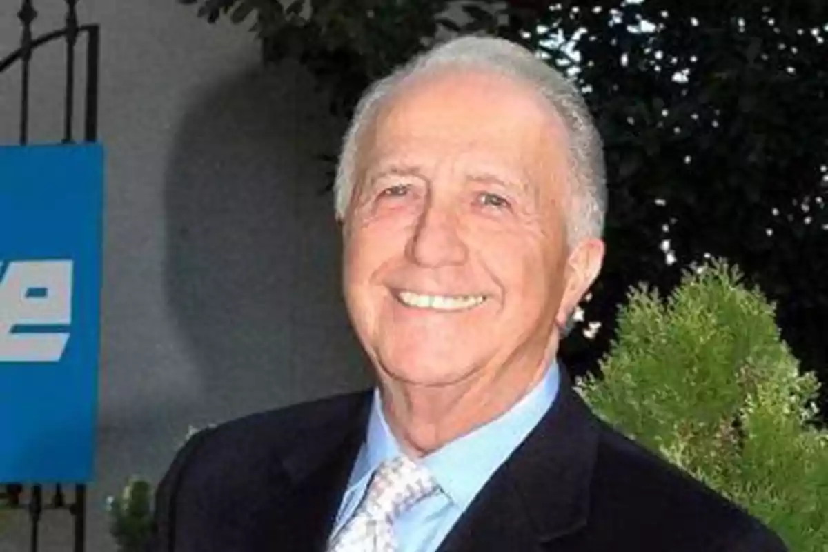 Un hombre mayor sonriendo, vestido con traje y corbata, en un entorno exterior con vegetación. Un hombre mayor sonriendo, vestido con traje y corbata, en un entorno exterior con vegetación.