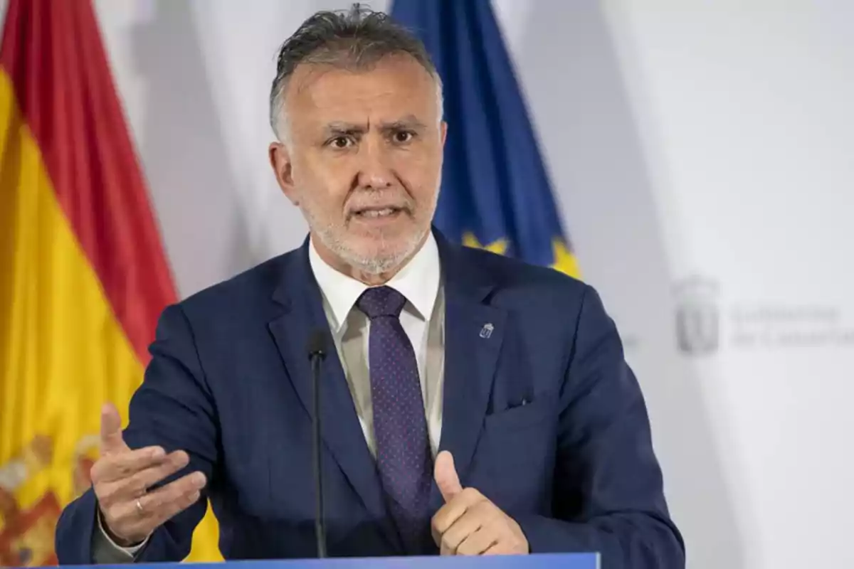 Hombre de traje azul hablando en una conferencia con banderas de España y la Unión Europea de fondo.