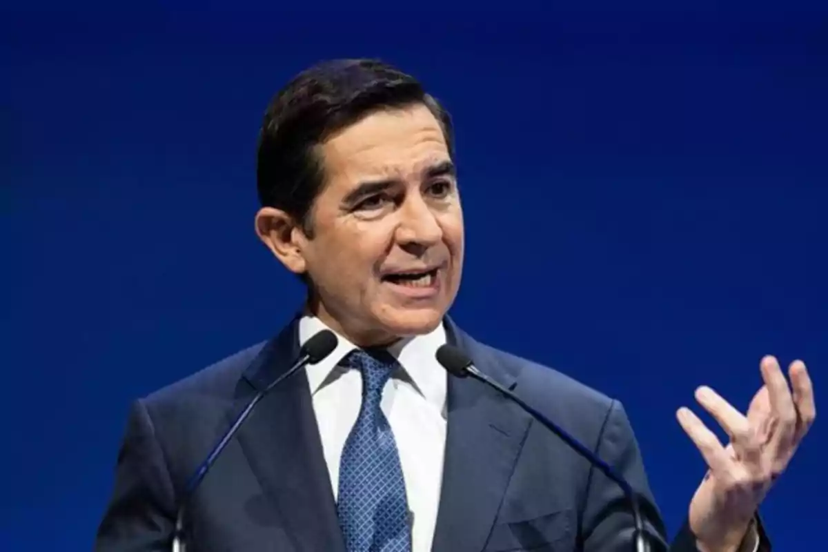 Hombre de traje y corbata hablando en público frente a dos micrófonos con fondo azul