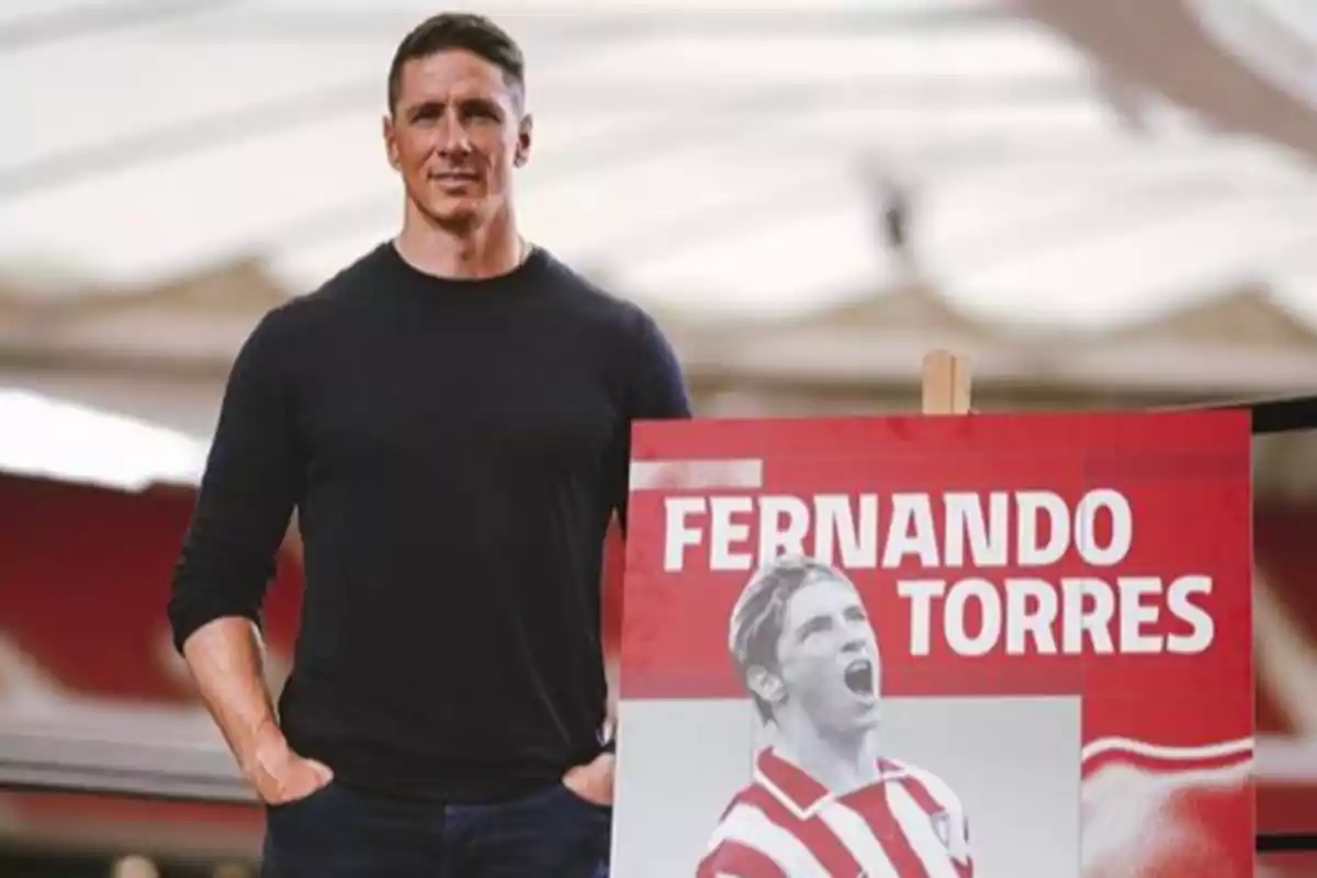 Hombre de pie junto a un cartel rojo con el nombre Fernando Torres y una imagen de un futbolista celebrando con la camiseta del Atlético de Madrid Hombre de pie junto a un cartel rojo con el nombre Fernando Torres y una imagen de un futbolista celebrando con la camiseta del Atlético de Madrid