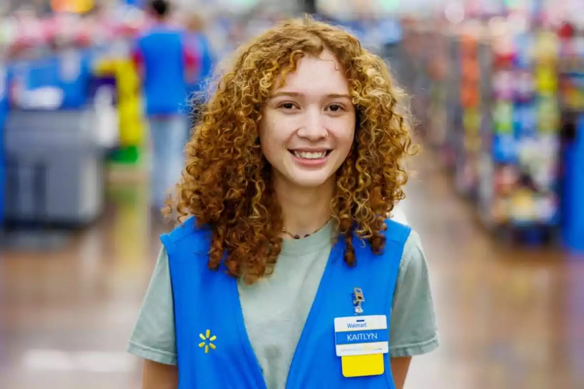 Una persona con cabello rizado y uniforme azul sonríe en el pasillo de una tienda Walmart. Una persona con cabello rizado y uniforme azul sonríe en el pasillo de una tienda Walmart.