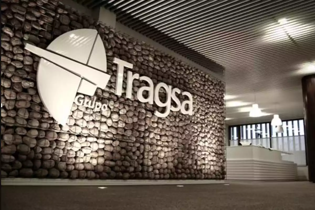 Logotipo de Grupo Tragsa en una pared de piedra dentro de una oficina moderna con techo de listones y lámparas colgantes.