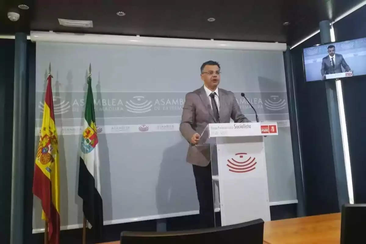 Un hombre de pie en un podio con el logotipo de la Asamblea de Extremadura, acompañado de las banderas de España y Extremadura, mientras habla en una conferencia de prensa. Un hombre de pie en un podio con el logotipo de la Asamblea de Extremadura, acompañado de las banderas de España y Extremadura, mientras habla en una conferencia de prensa.