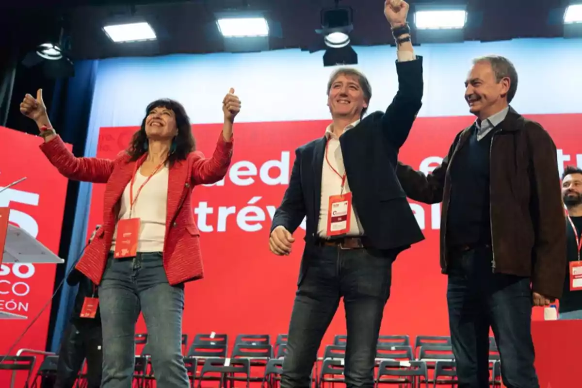 Tres personas sonríen y celebran en un escenario con fondo rojo y sillas vacías, usando gafetes colgados al cuello Tres personas sonríen y celebran en un escenario con fondo rojo y sillas vacías, usando gafetes colgados al cuello