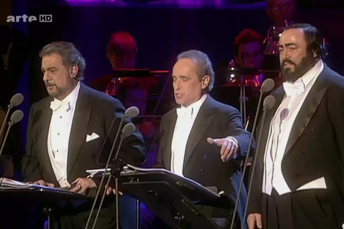 Tres cantantes de ópera vestidos de gala interpretan en un escenario con micrófonos y una orquesta de fondo. Tres cantantes de ópera vestidos de gala interpretan en un escenario con micrófonos y una orquesta de fondo.