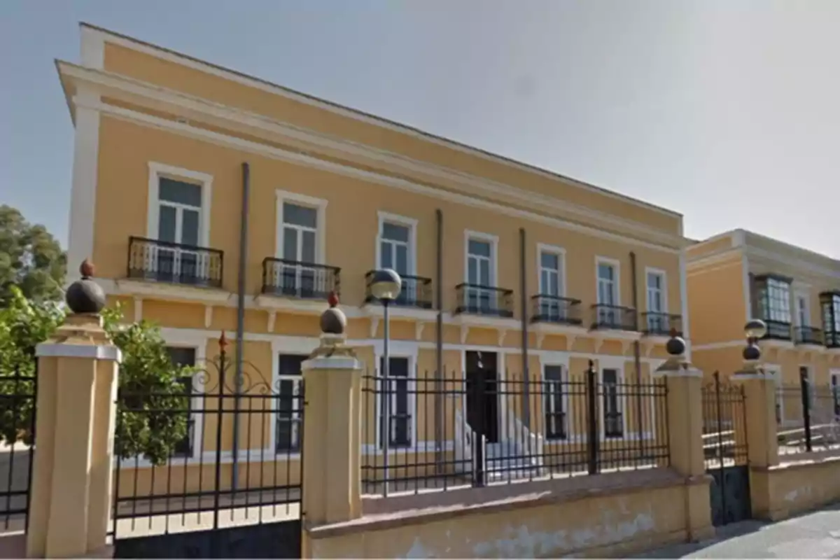 Edificio de dos pisos de color amarillo con balcones y ventanas blancas, rodeado por una reja de metal negro y pilares amarillos.