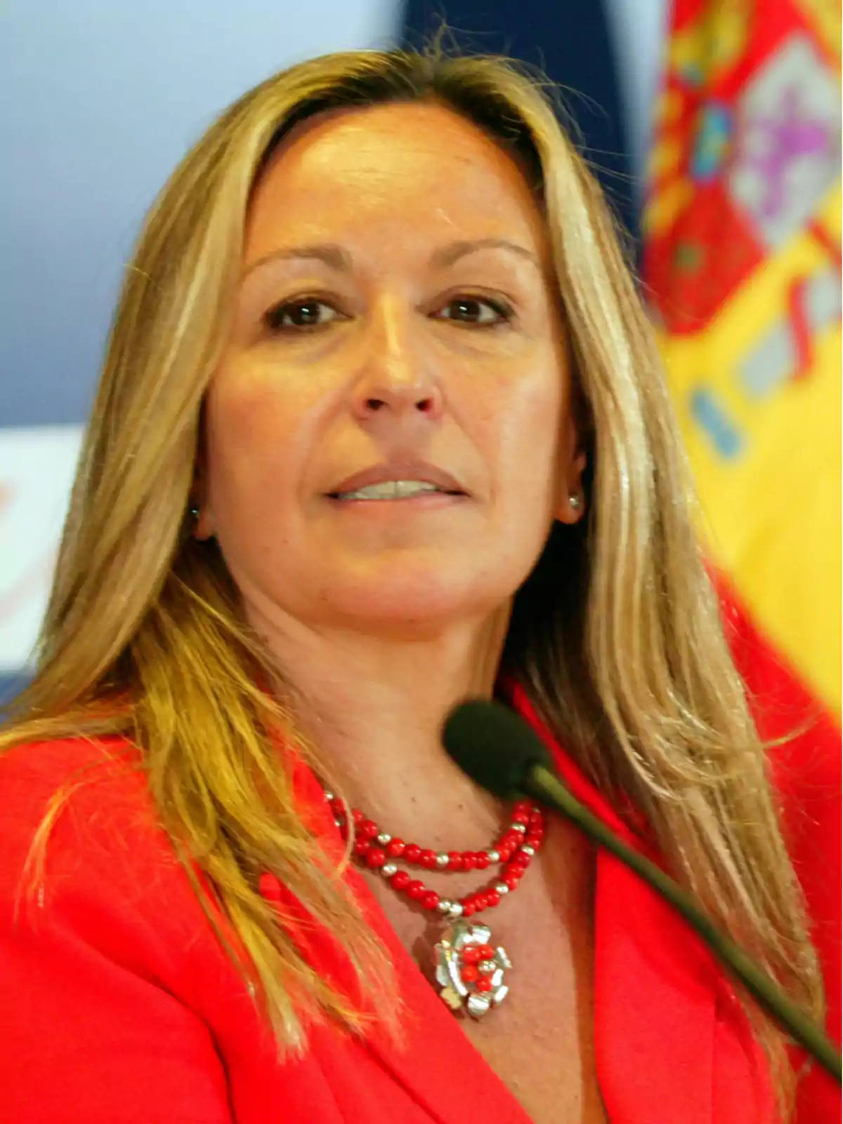 Mujer con cabello rubio y chaqueta roja hablando frente a un micrófono con una bandera de España en el fondo.