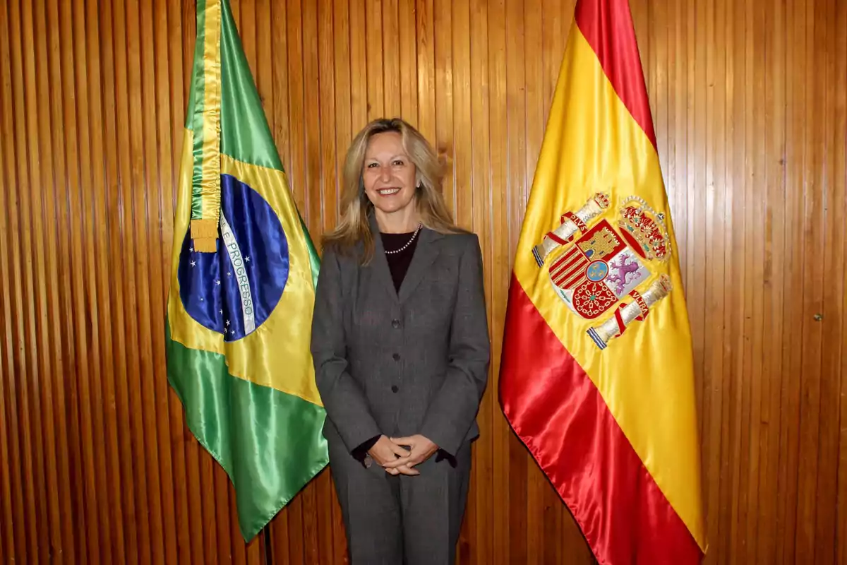Una mujer de pie entre las banderas de Brasil y España, con una pared de madera de fondo.