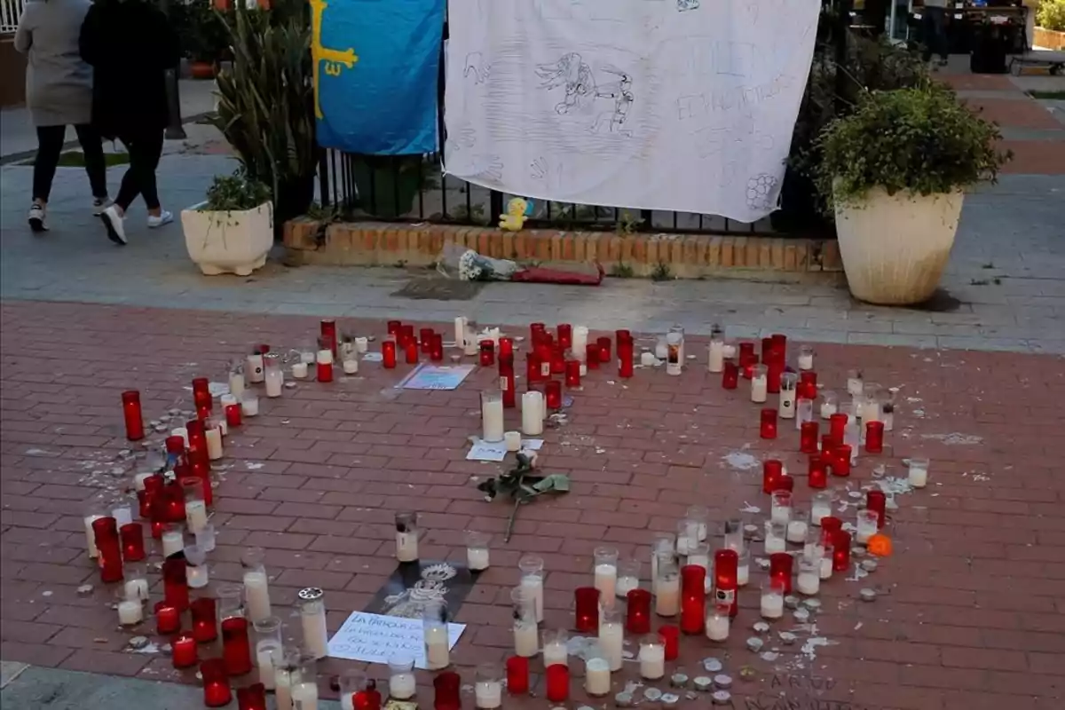 Un altar con velas rojas y blancas dispuestas en un círculo sobre el suelo de ladrillo, con una bandera azul y un cartel blanco con mensajes y dibujos colgados en una cerca, y dos personas caminando al fondo.