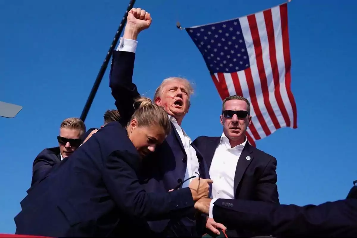 Un grupo de personas, algunas con gafas de sol, rodean a un hombre que levanta el puño, con una bandera de Estados Unidos ondeando en el fondo.