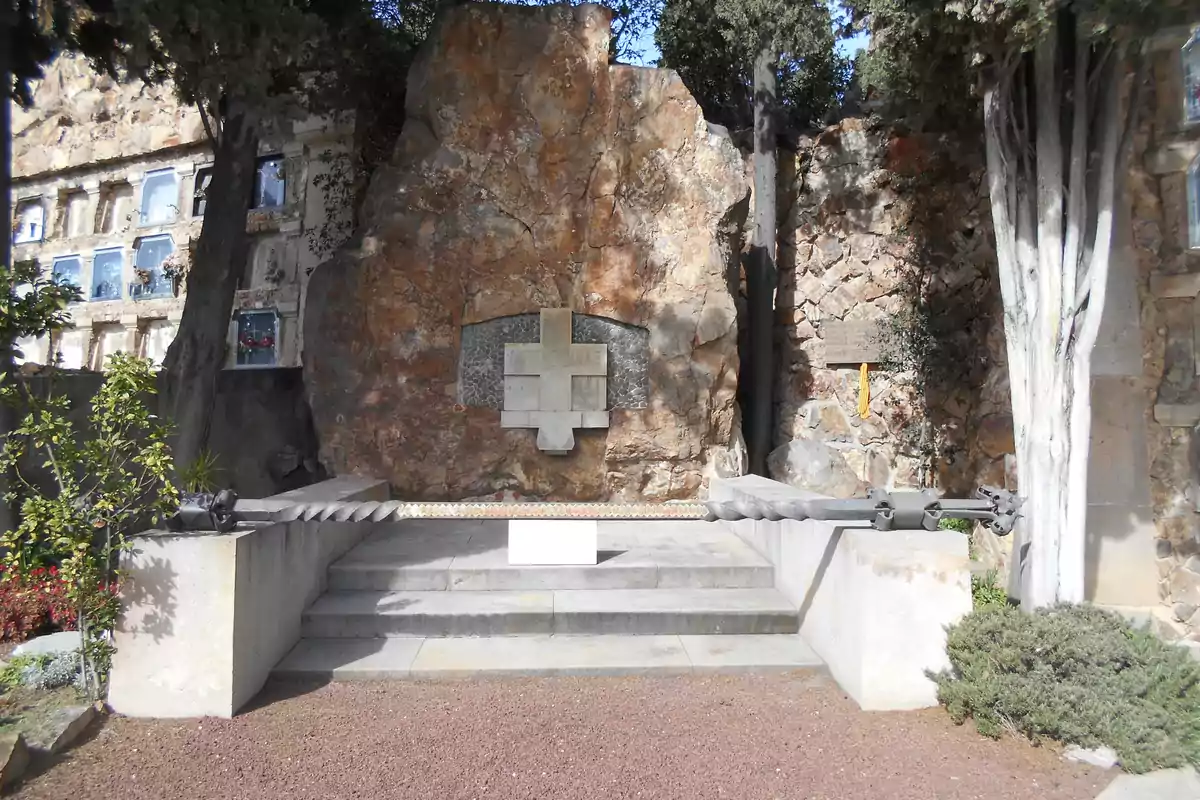 Una tumba en un cementerio con una gran roca de fondo y una cruz en el centro rodeada de árboles y nichos en la pared lateral.