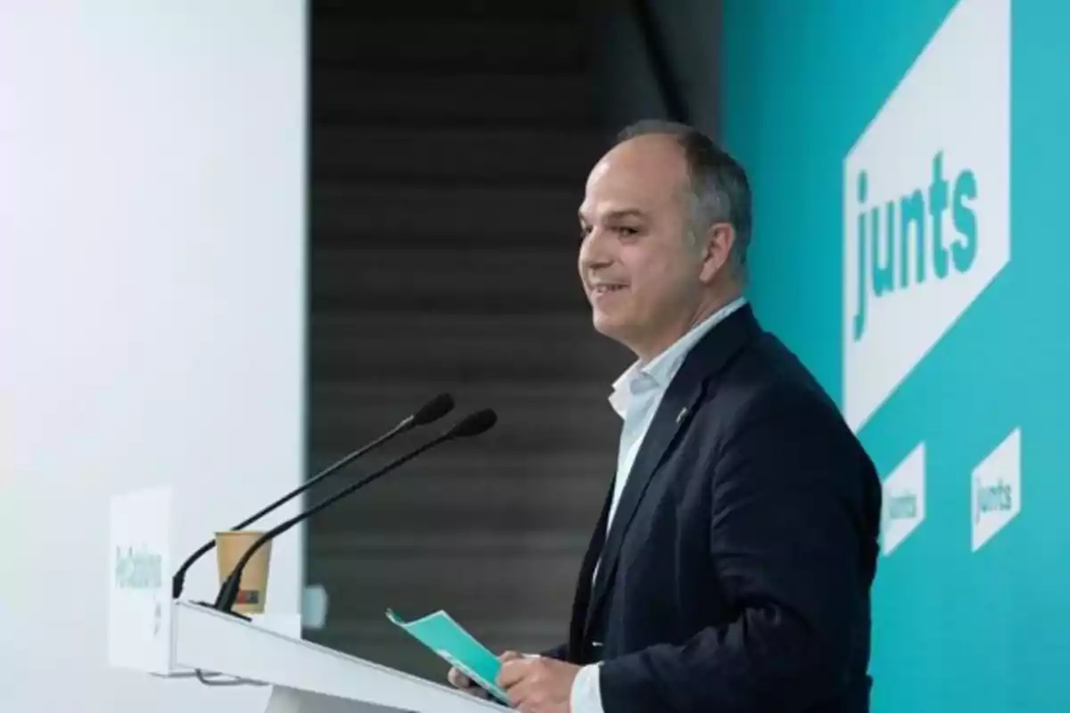Hombre de traje hablando en un atril con fondo azul y el logo de junts Hombre de traje hablando en un atril con fondo azul y el logo de junts