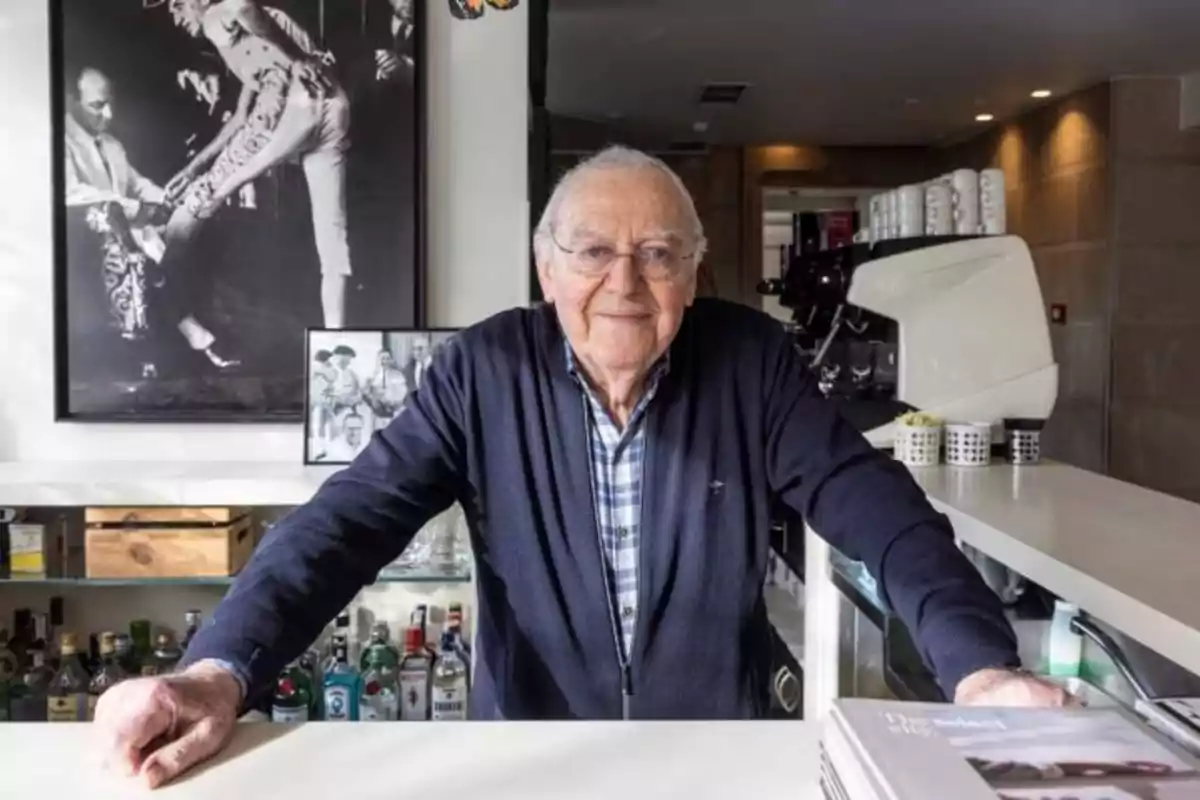 Un hombre mayor con gafas y suéter oscuro está de pie detrás de una barra, con botellas de licor en el estante detrás de él y una máquina de café a su lado; en la pared hay una fotografía en blanco y negro de un torero.