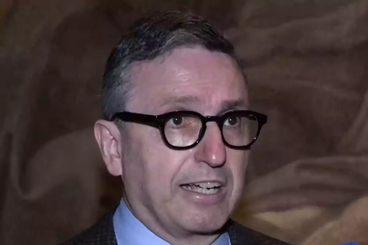 Un hombre con gafas y cabello corto está hablando frente a un fondo oscuro. Un hombre con gafas y cabello corto está hablando frente a un fondo oscuro.