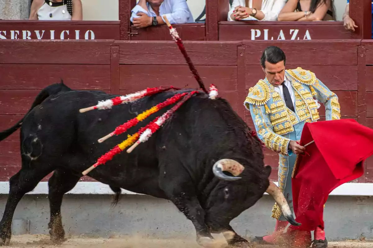 Un torero vestido con traje de luces realiza una faena con un toro en una plaza de toros, mientras el público observa desde las gradas.