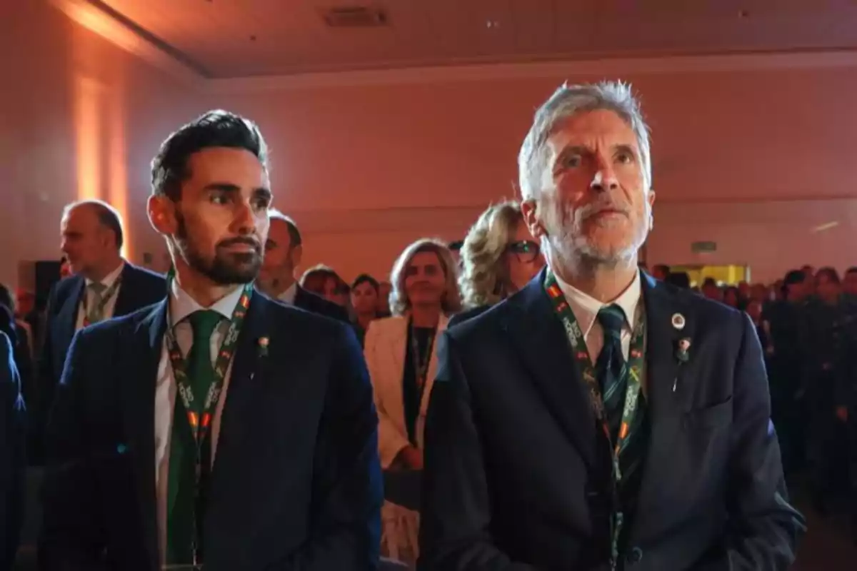 Dos hombres con traje y corbata están sentados en un evento formal junto a otras personas en un salón iluminado con luces cálidas Dos hombres con traje y corbata están sentados en un evento formal junto a otras personas en un salón iluminado con luces cálidas