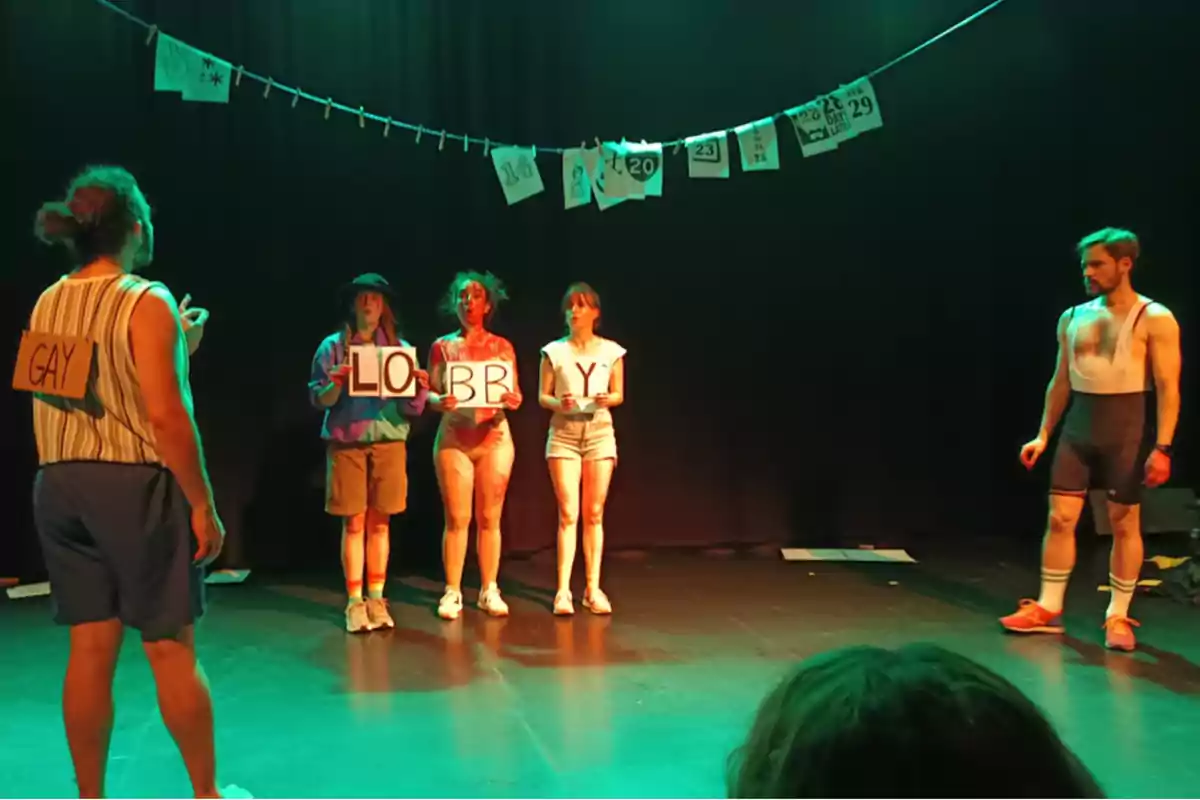 Cinco personas en un escenario teatral con iluminación verde, tres de ellas sostienen carteles que forman la frase lobby y una persona lleva un cartel en la espalda que dice gay mientras otra observa desde la derecha