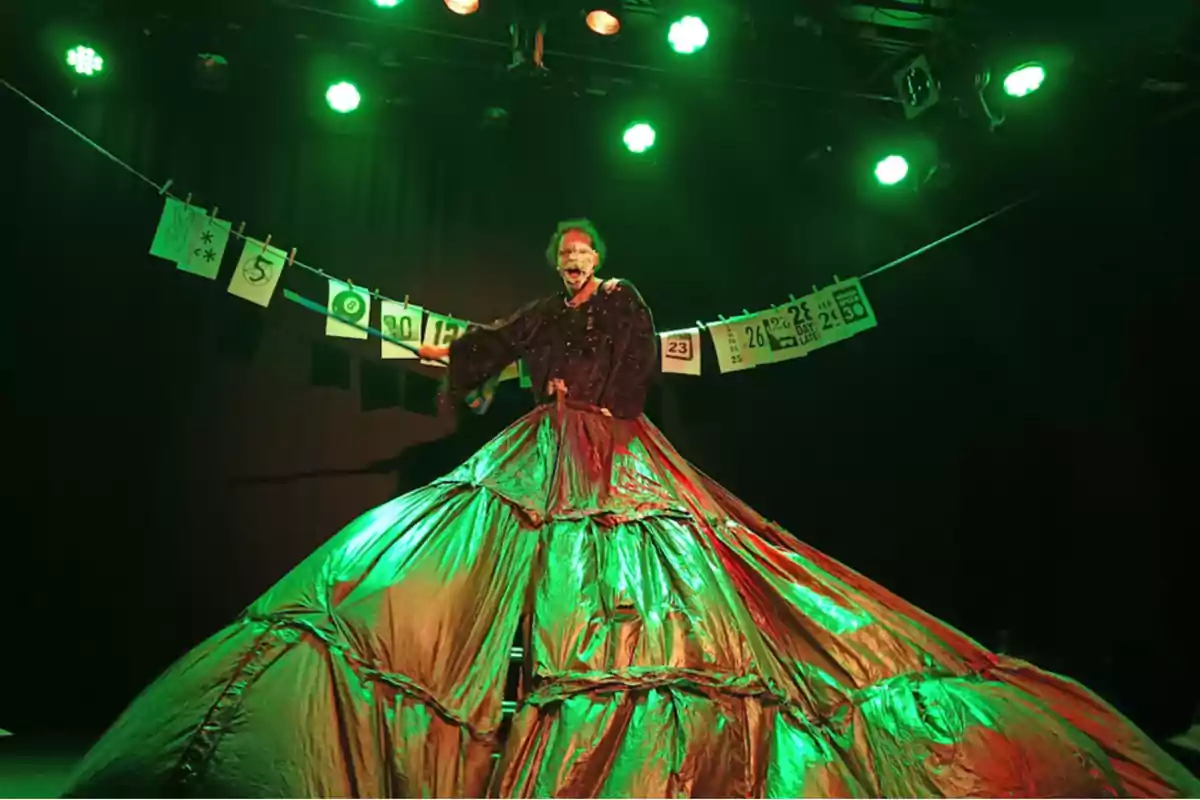 Persona maquillada con vestuario teatral voluminoso bajo luces verdes en un escenario con papeles colgados en el fondo Persona maquillada con vestuario teatral voluminoso bajo luces verdes en un escenario con papeles colgados en el fondo