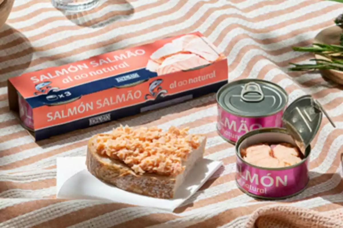 Una lata de salmón al natural de Mercadona abierta junto a una rebanada de pan con salmón desmenuzado sobre un mantel a rayas. Una lata de salmón al natural de Mercadona abierta junto a una rebanada de pan con salmón desmenuzado sobre un mantel a rayas.