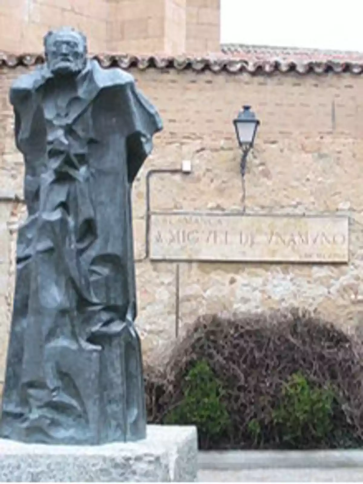 Escultura de un hombre con capa en un entorno urbano con una pared de ladrillo y una farola.