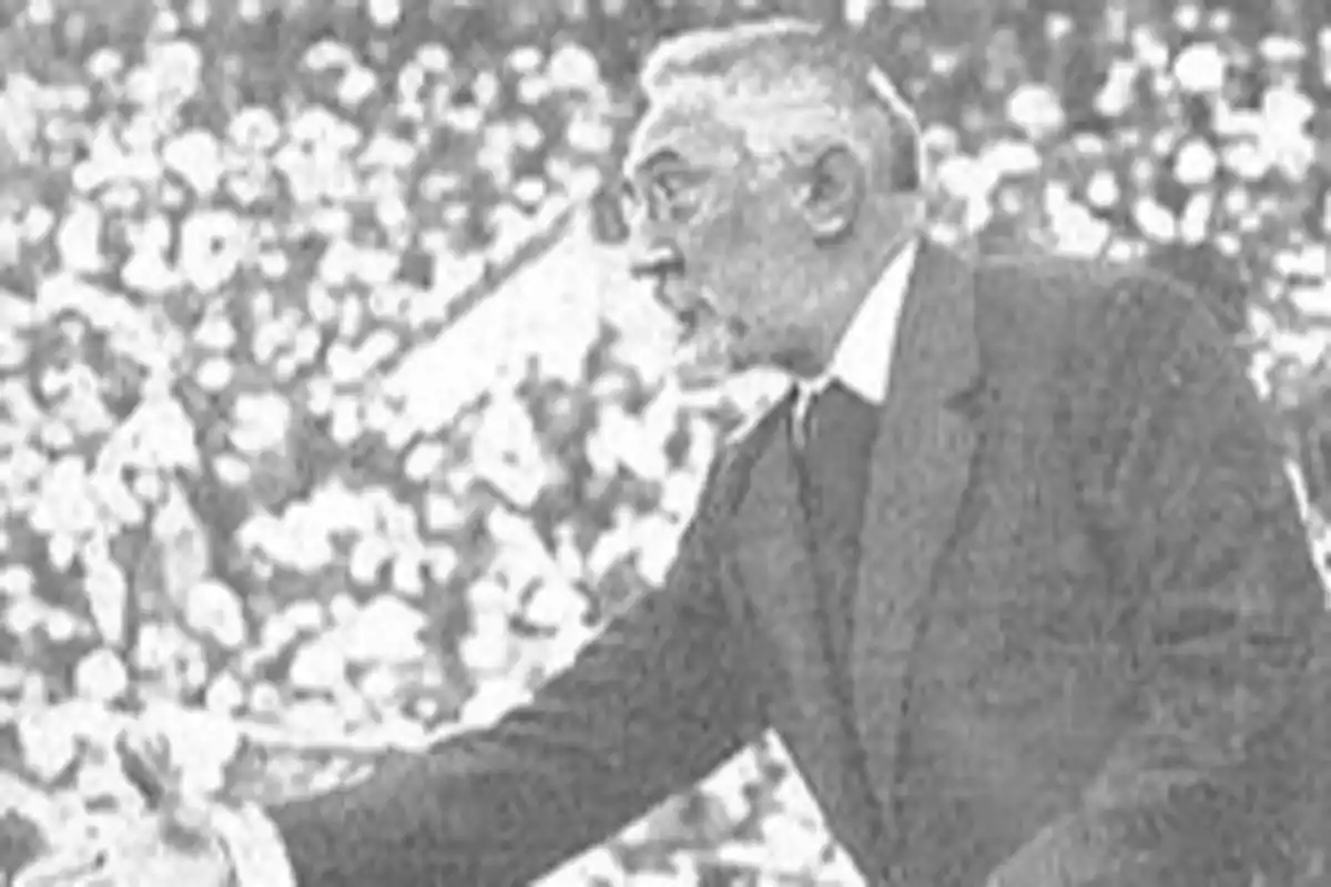 Un hombre mayor con barba y gafas, vestido con traje, gesticulando en un entorno con muchas personas.