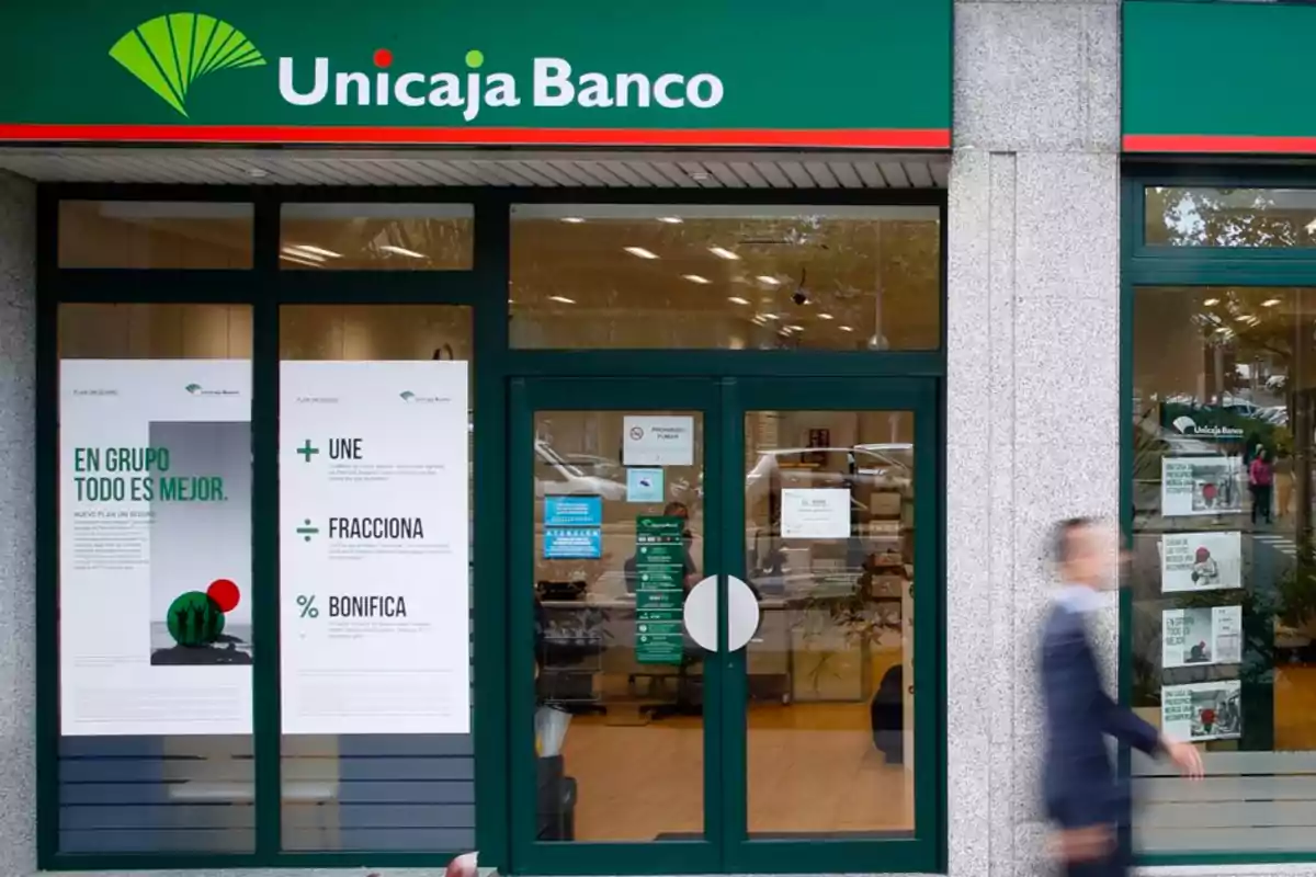 Fachada de una sucursal de Unicaja Banco con carteles promocionales en las ventanas y una persona caminando frente a la entrada.