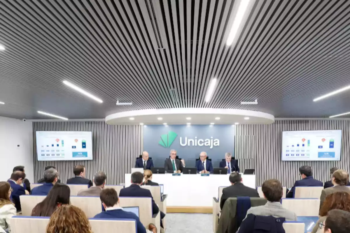 Personas en una conferencia de prensa en una sala moderna con el logo de Unicaja en la pared y pantallas mostrando gráficos.