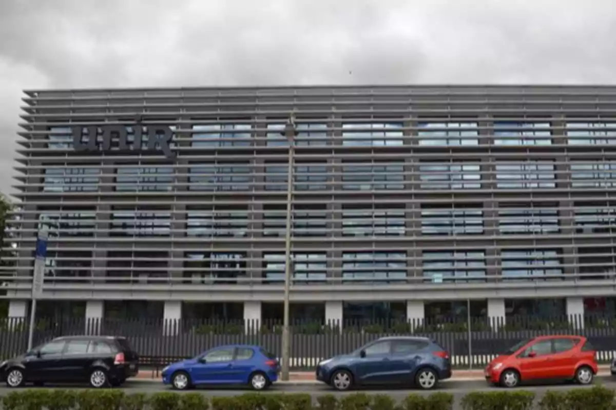 Edificio moderno con fachada de lamas horizontales y varios coches estacionados en la calle frente a él.