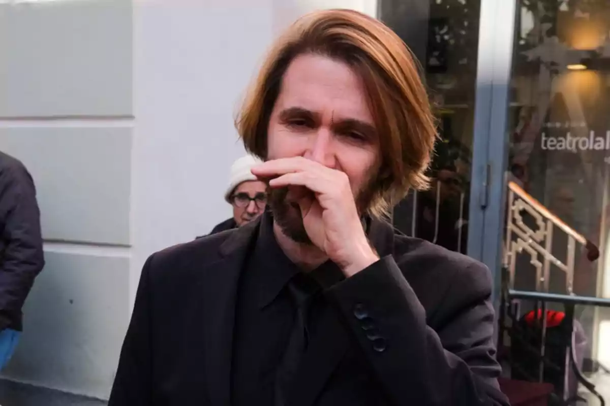 Un hombre con barba y cabello largo, vestido con un traje negro, se cubre la boca con la mano mientras está de pie frente a un edificio con un letrero que dice 