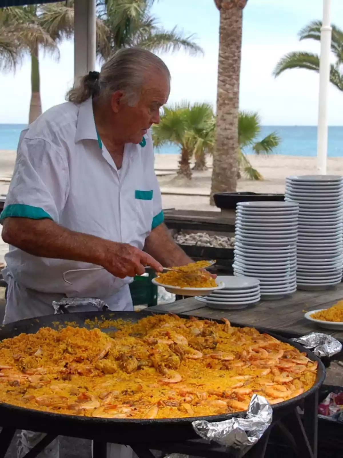 Un hombre sirviendo paella en un plato junto a una gran paellera en un entorno al aire libre con palmeras y el mar de fondo.
